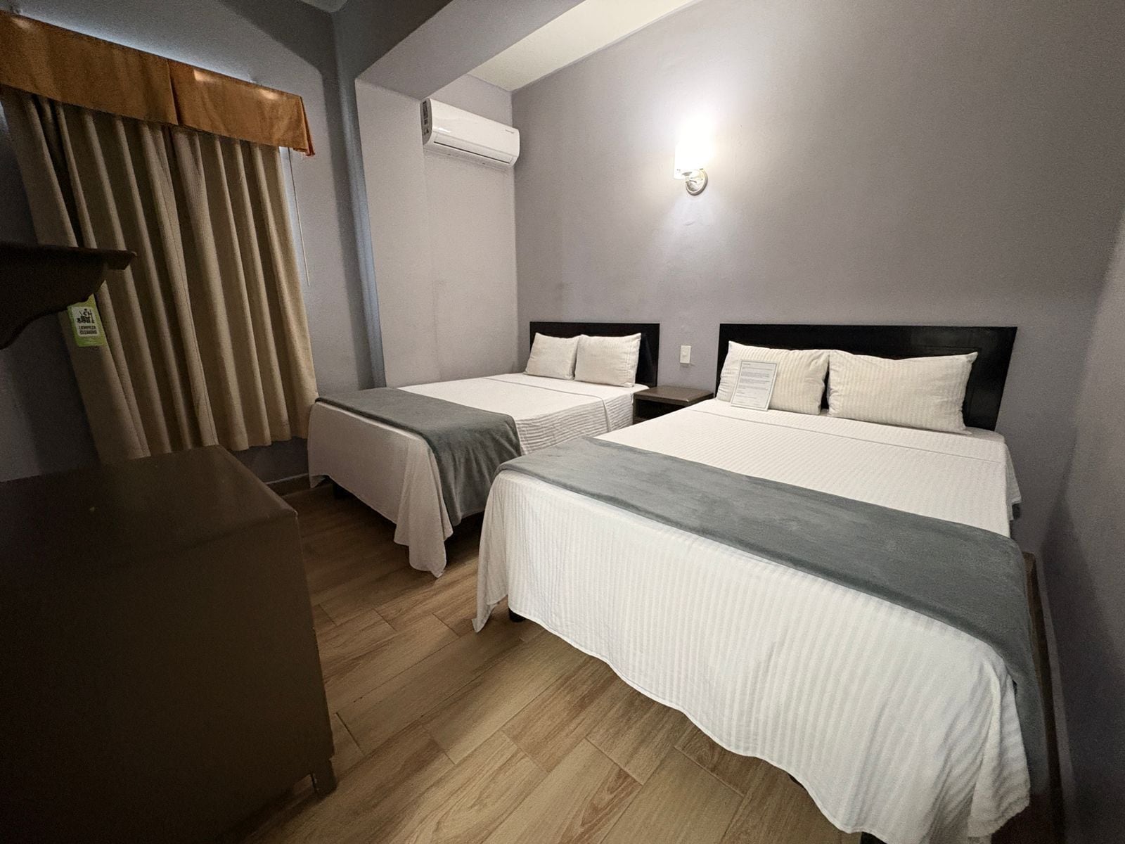 A hotel room with two single beds, a wooden floor, an air conditioning unit, and a small desk.