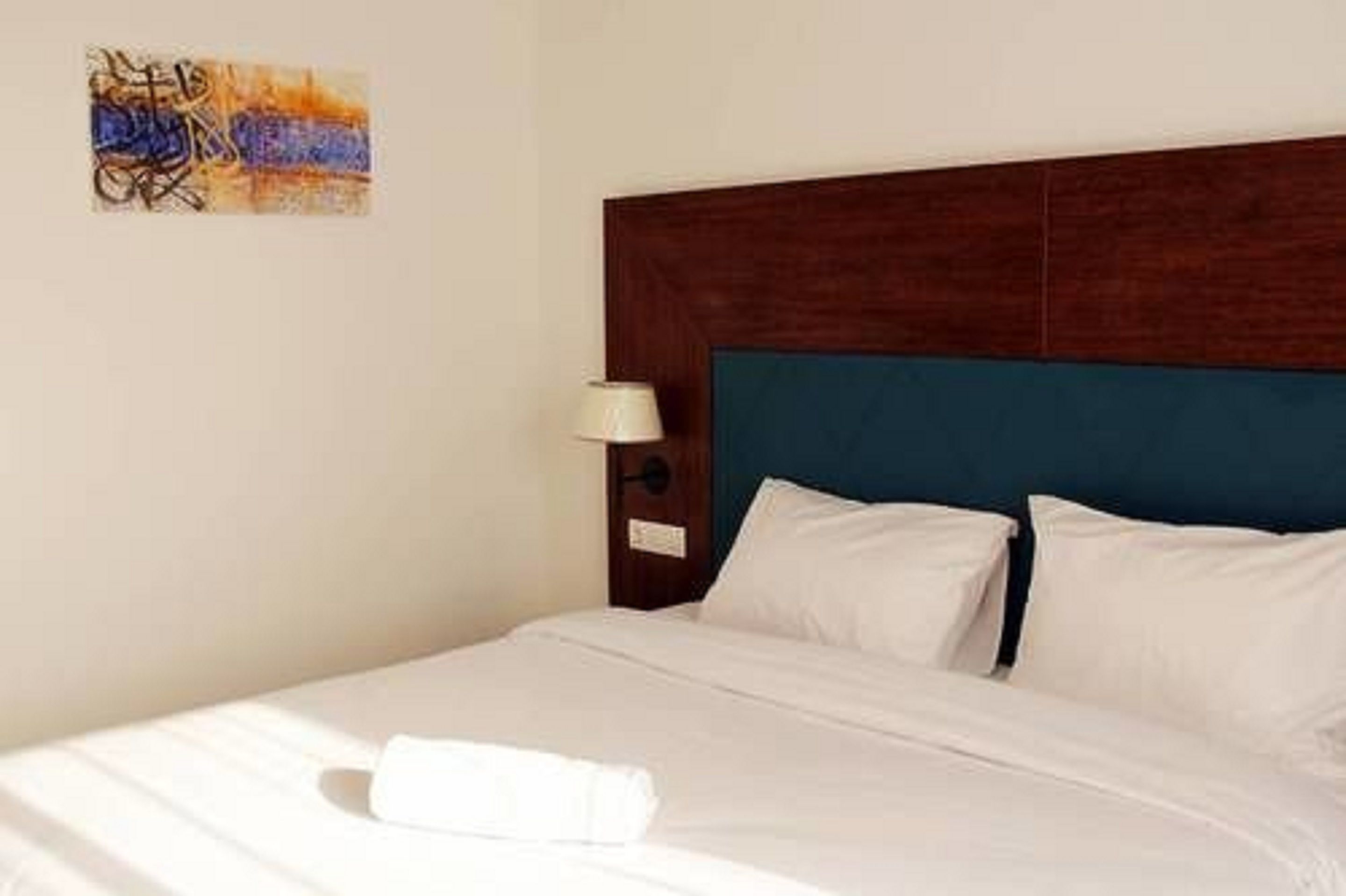 A neatly made bed with white linens and pillows, a wooden headboard, and a wall-mounted lamp.