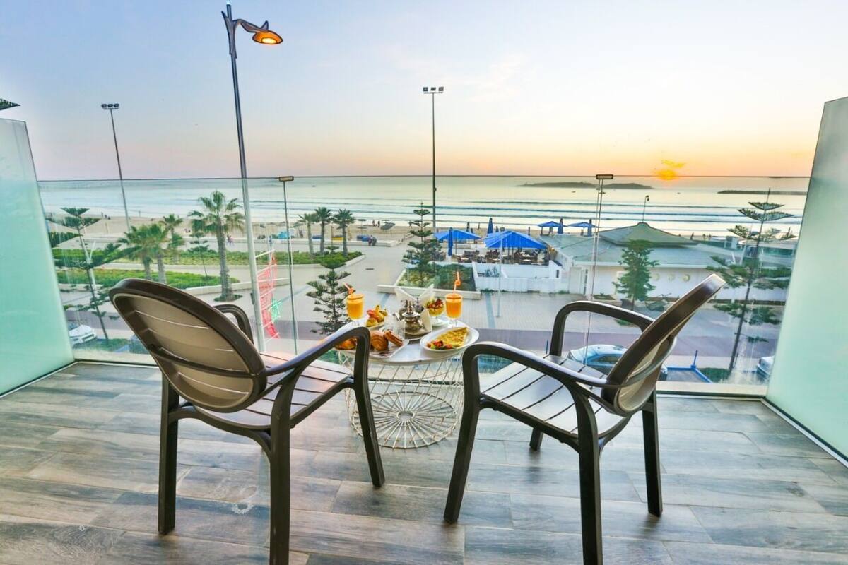 Un balcon avec une table préparée pour le petit-déjeuner, d’où l’on peut admirer une plage et un coucher de soleil.