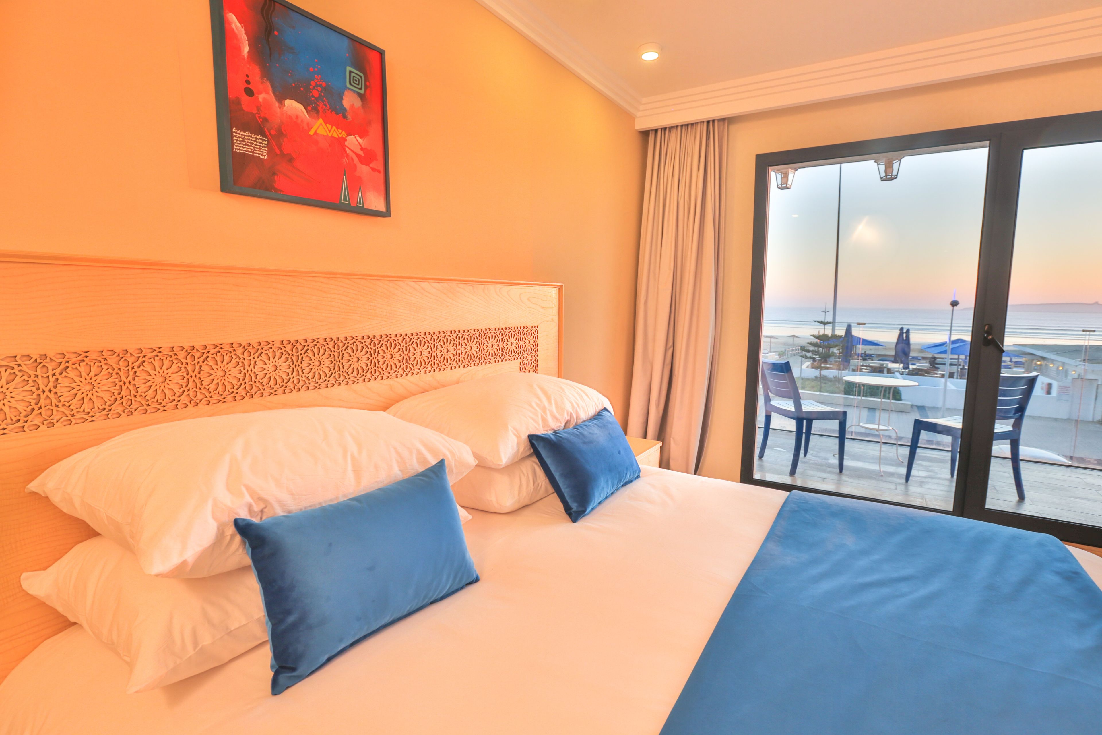 A hotel room with a large bed, two blue pillows, a headboard with a decorative pattern, a painting on the wall, and a view of the beach through a large window.