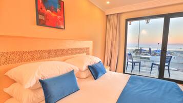 A hotel room with a large bed, two blue pillows, a headboard with a decorative pattern, a painting on the wall, and a view of the beach through a large window.