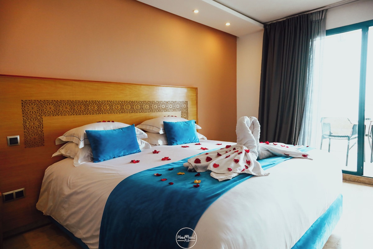Ein Hotelzimmer mit einem großen Bett, blau-weißer Bettwäsche und Blick ins Freie durch ein Fenster.