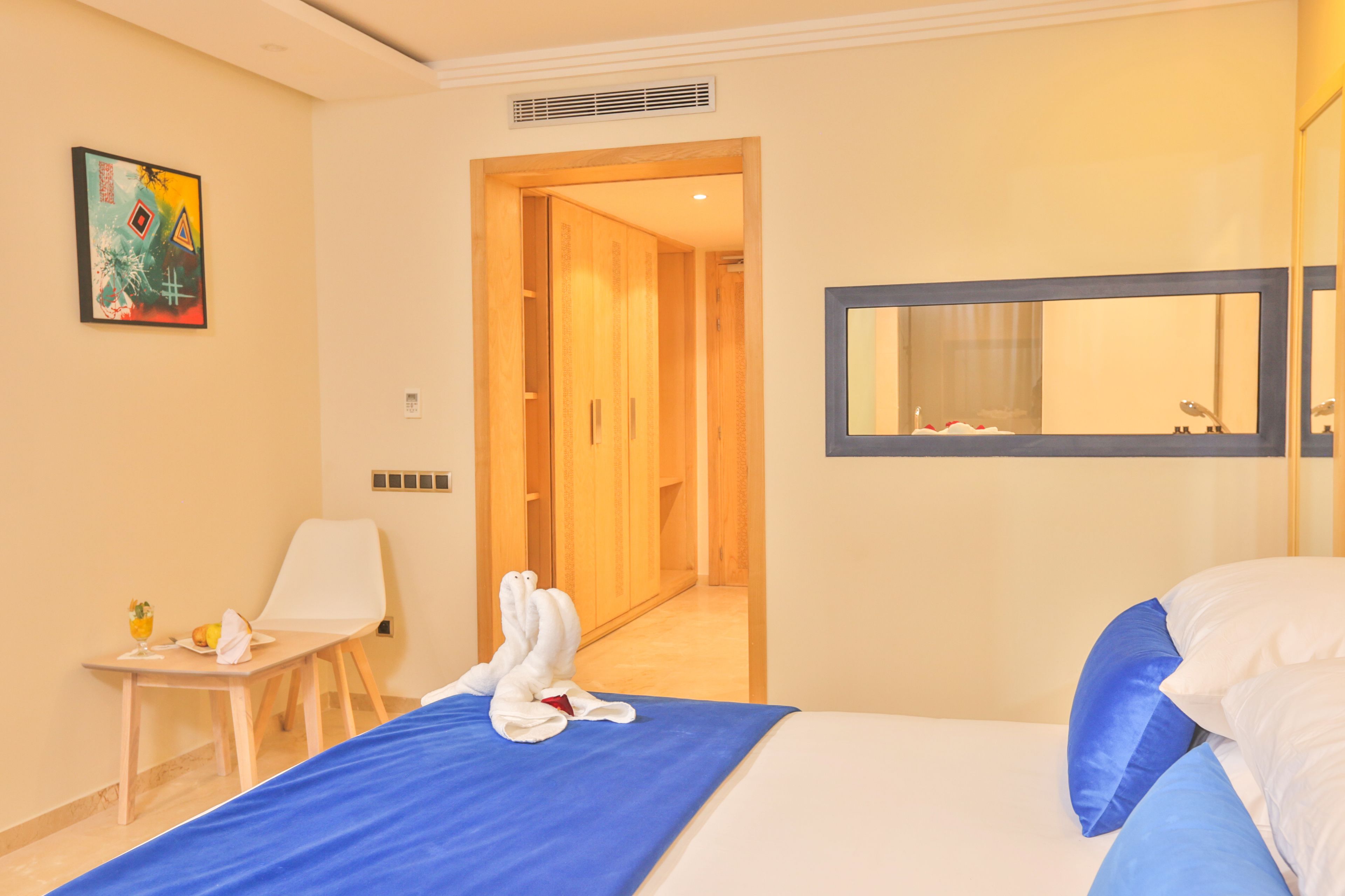 Una habitación de hotel con una cama, un edredón azul, una silla blanca, una mesita con una toalla y un vaso de agua, y un cuadro en la pared.