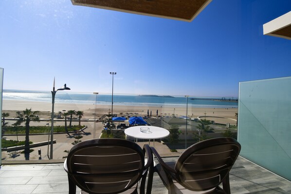 Ein Balkon mit zwei Stühlen und einem Tisch mit Blick auf einen Strand, an dem Palmen und Sonnenschirme stehen.