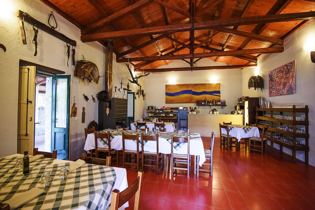 Un comedor rústico con mesas y sillas de madera, una cocina al fondo y un cuadro grande en la pared.