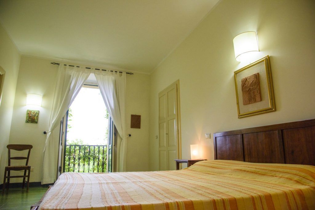 Ein Hotelzimmer mit Bett, Stuhl, einem Balkon mit Vorhängen, einer Tür und einer Wandleuchte.