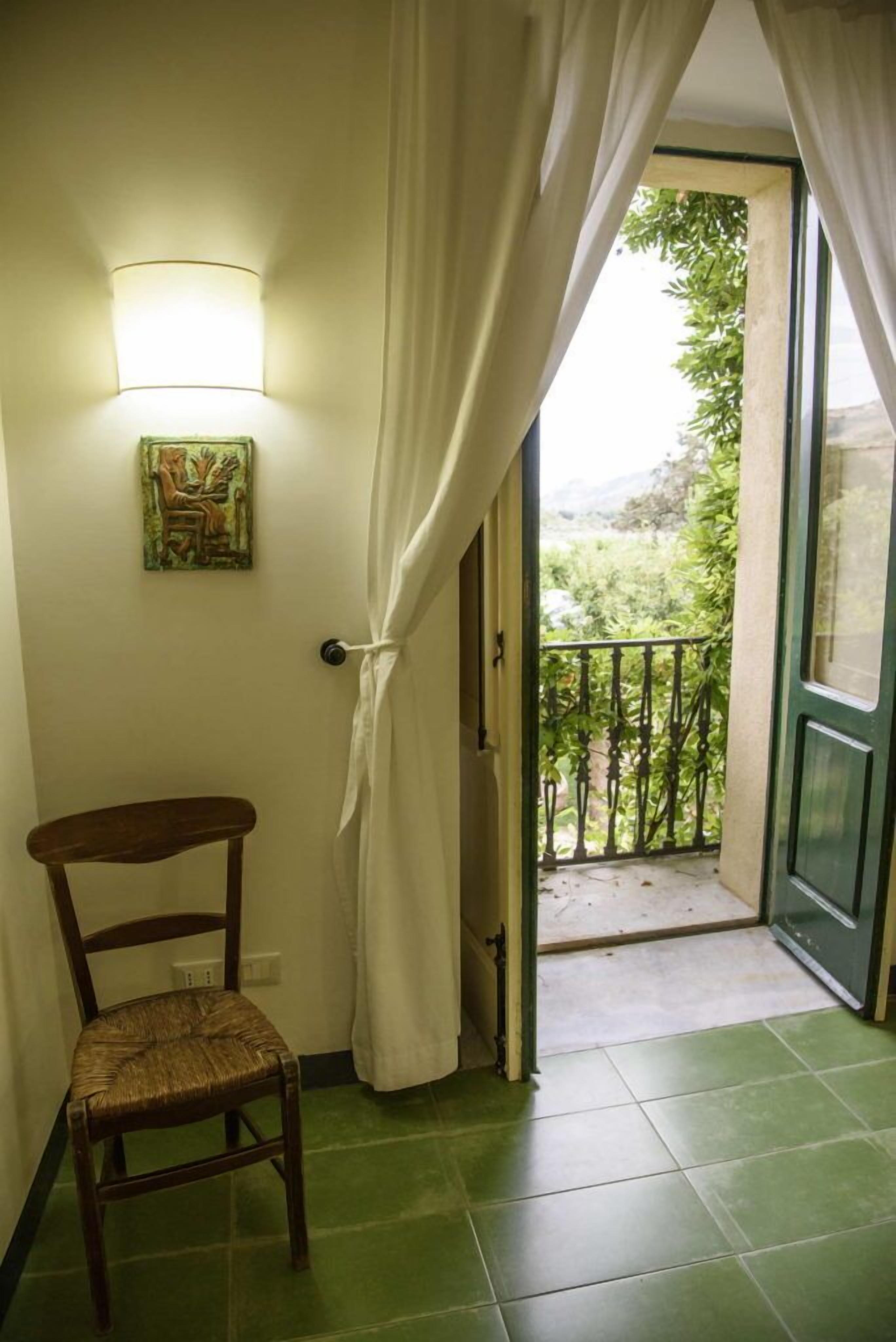 A room with a chair, a wall-mounted light, a door leading to a balcony, and a view of greenery outside.