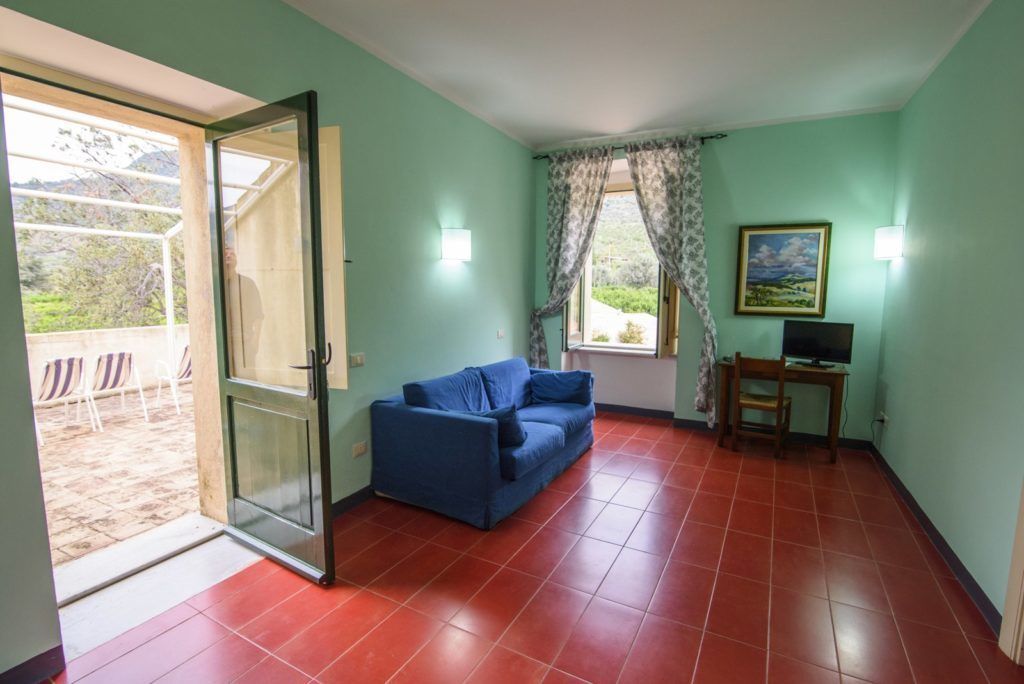 A room with a blue sofa, a small table with a TV, a window with curtains, and a door leading to an outdoor area with chairs.