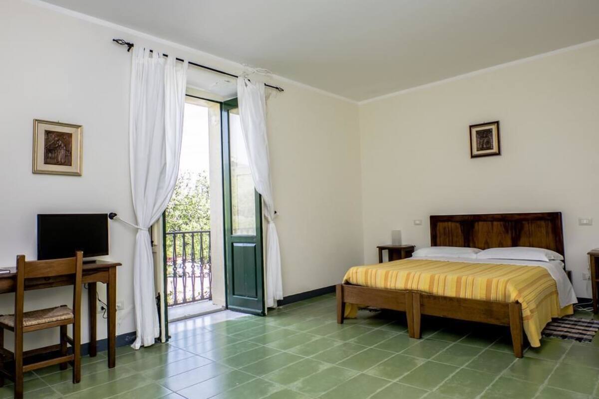 Ein Schlafzimmer mit Bett, Schreibtisch mit Stuhl, Fernseher und einer Tür, die auf einen Balkon führt.