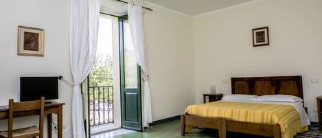 A bedroom with a bed, a desk with a chair, a television, and a door leading to a balcony.
