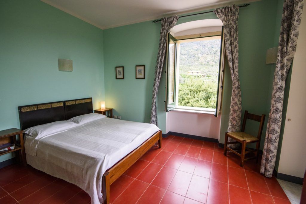 Quarto com cama, uma cadeira, janela com cortinas e vista para o verde lá fora.