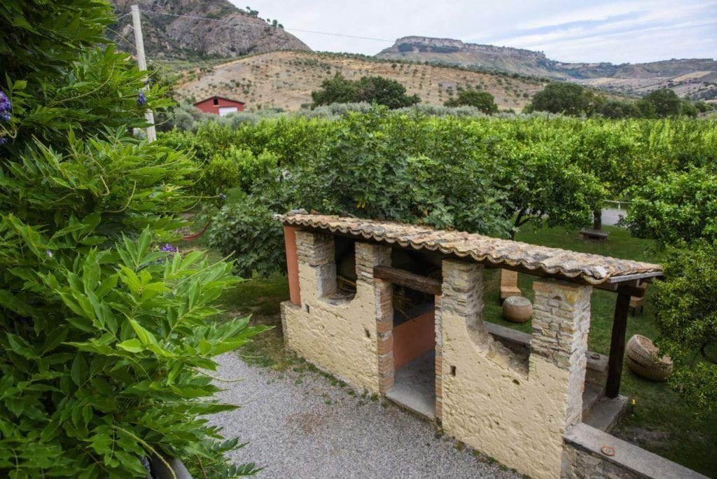 A rustic stone structure with a tiled roof, surrounded by lush greenery and vineyards in a hilly landscape.