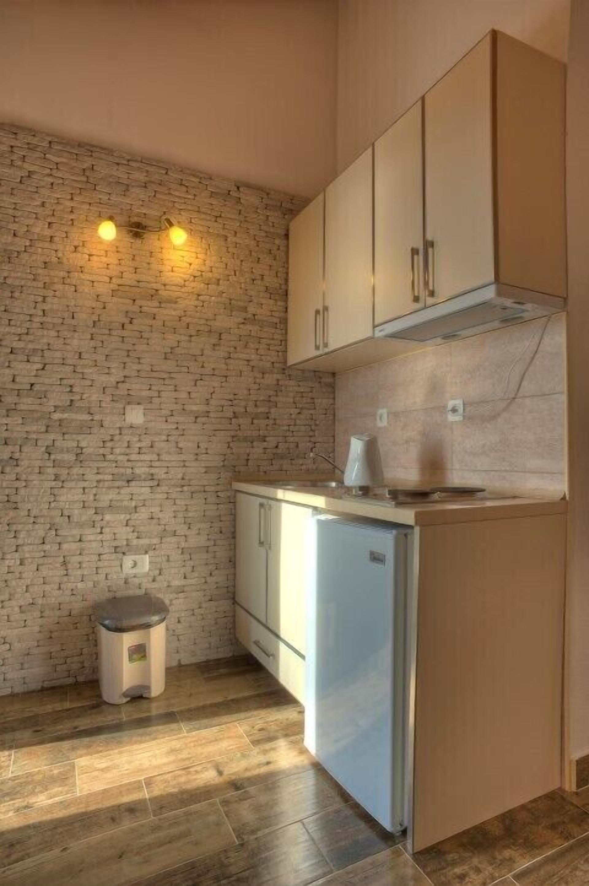 Cocina compacta con pared de ladrillo, armarios de madera, un pequeño cubo de basura y un refrigerador.