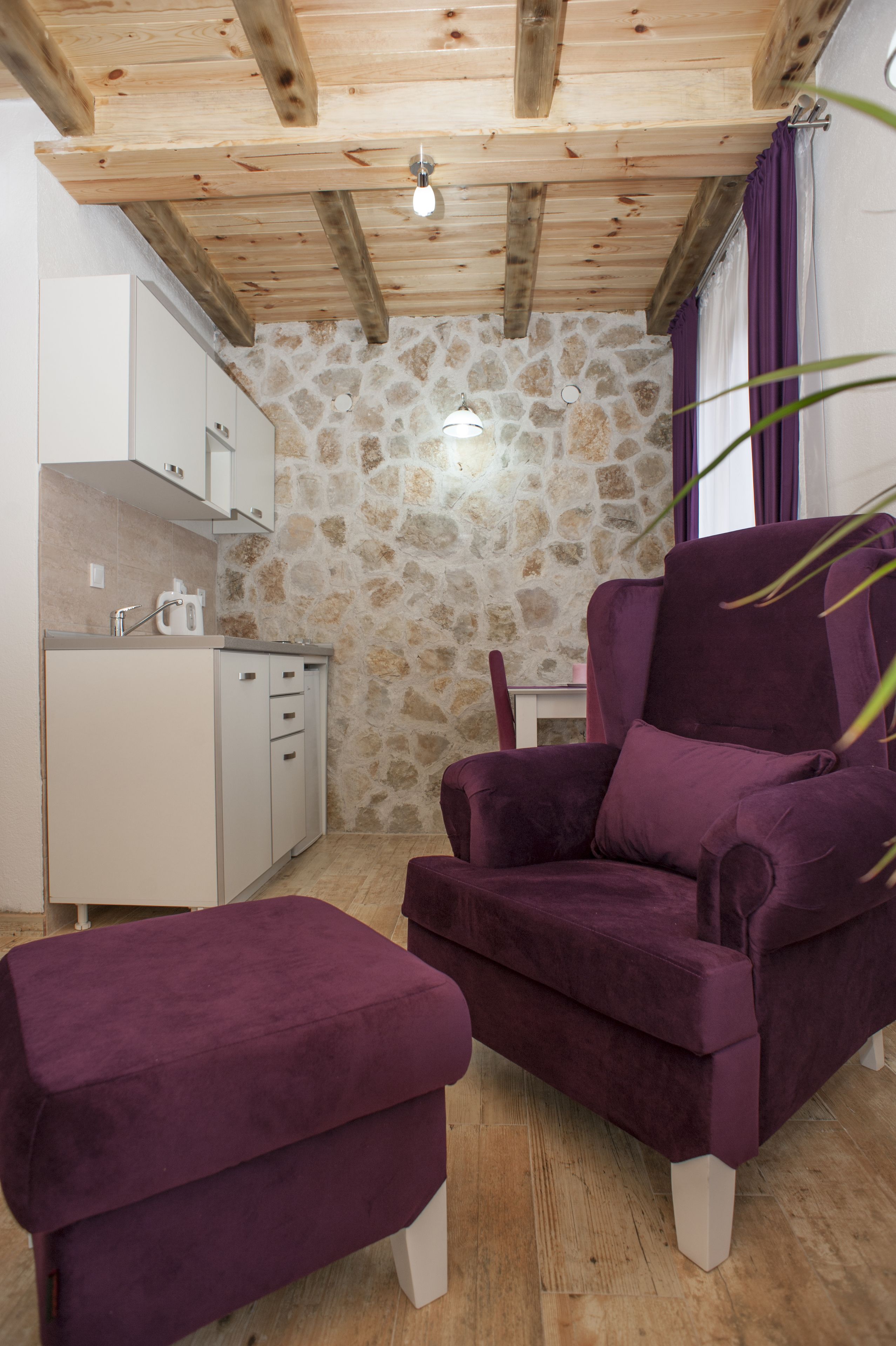 A room with a stone wall, wooden ceiling beams, a purple armchair, a matching ottoman, and a small kitchen area.