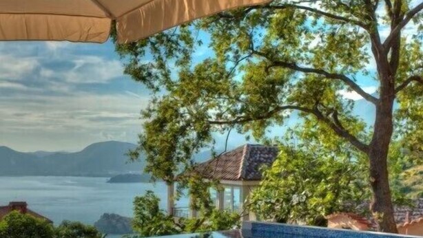 Piscina infinita con una tumbona de madera y sombrilla, rodeada de árboles y vista al mar.