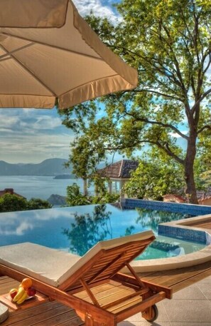 Piscina infinita con una tumbona de madera y sombrilla, rodeada de árboles y vista al mar.