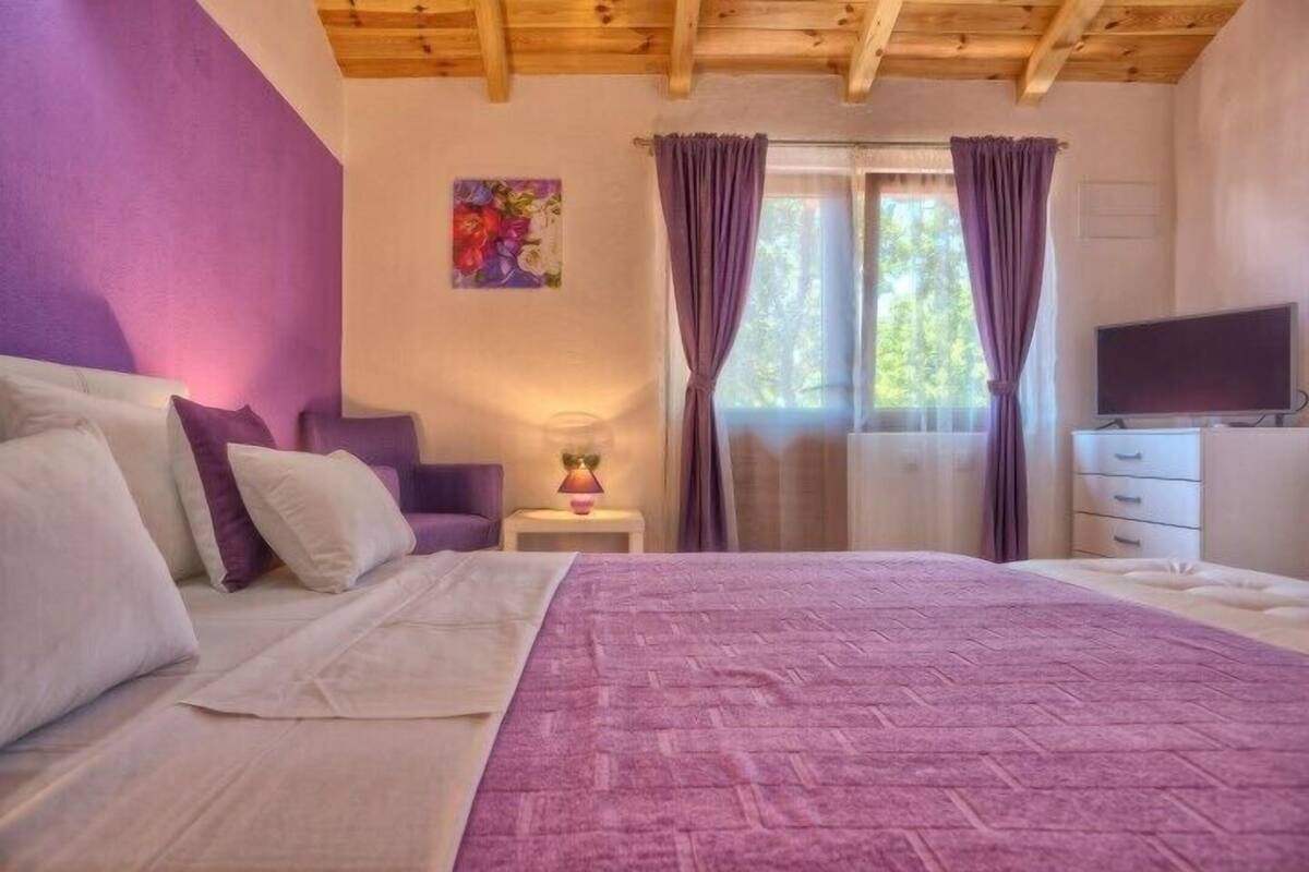 A bedroom with a bed, a nightstand, a lamp, a TV, and a window with curtains.