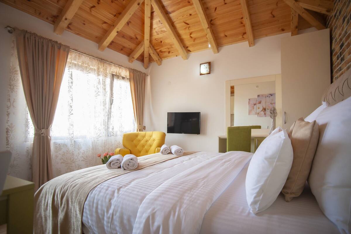 A hotel room with a wooden ceiling, a bed with white linens, a yellow armchair, and a flat-screen TV mounted on the wall.