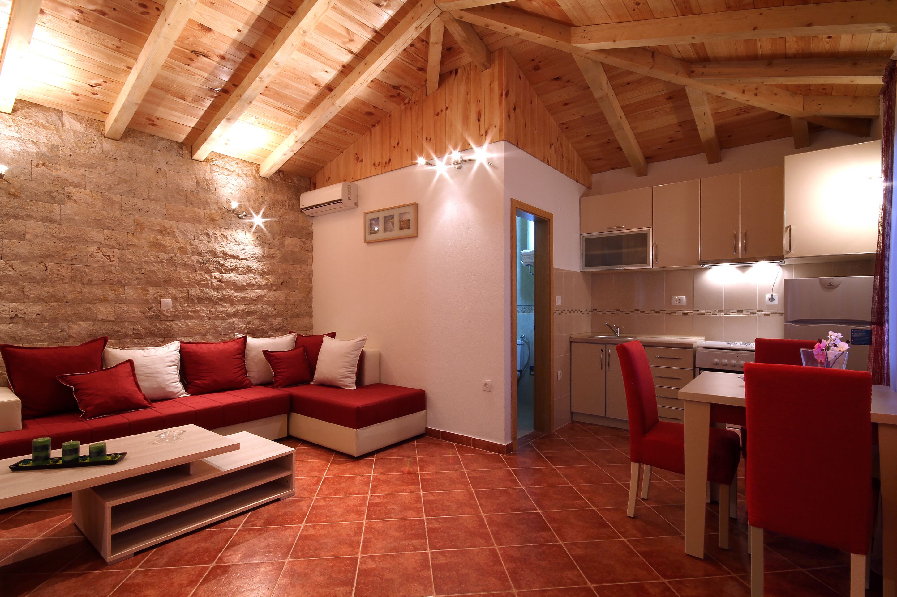 Una sala de estar acogedora con un sofá rojo, techo de madera y una zona de cocina con armarios blancos y sillas rojas.