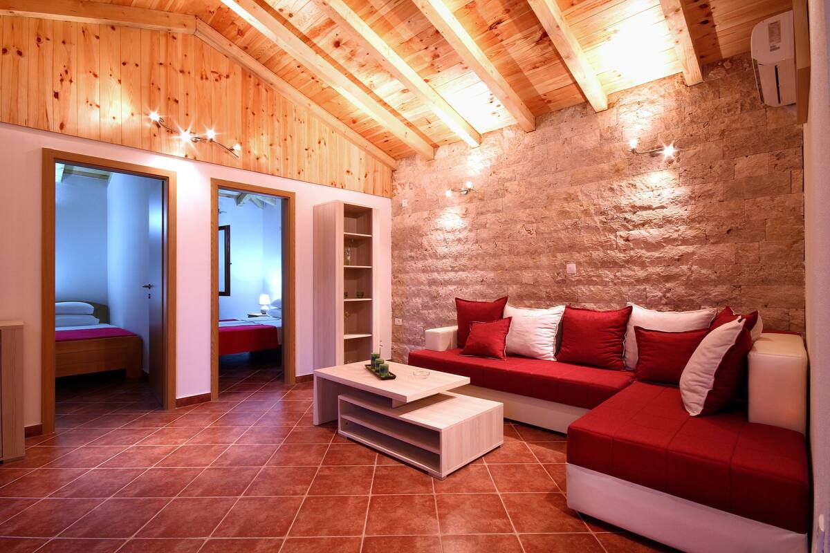 A cozy room with a wooden ceiling, a stone wall, a red sofa, and a bed with a red blanket.