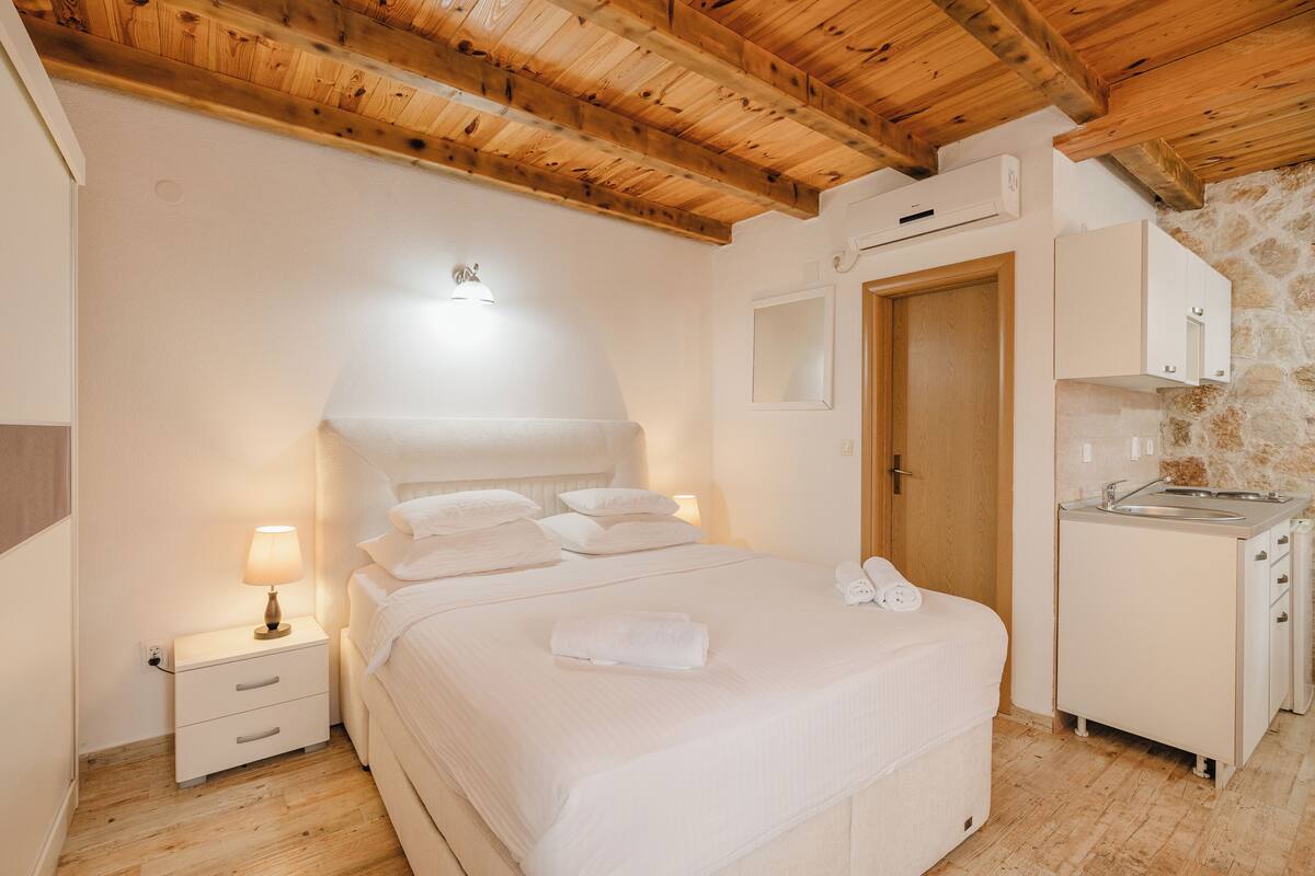 A bedroom with a wooden ceiling, a bed with white linens, a nightstand with a lamp, a wooden door, and a kitchenette with a sink.