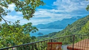 Terraza de madera con una mesa redonda y dos sillas con cojines, con vistas a un paisaje montañoso y un cuerpo de agua.