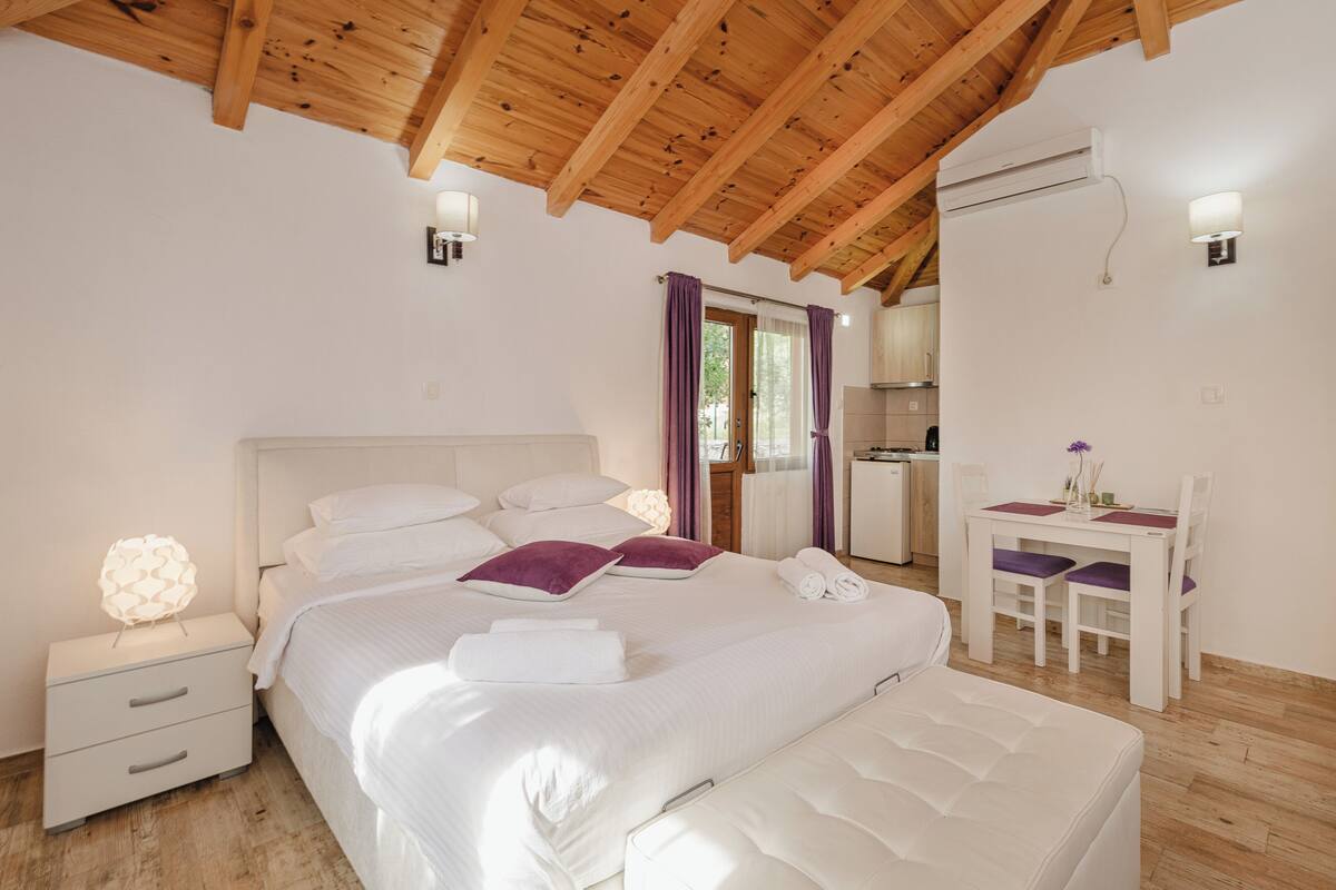 A bedroom with a wooden ceiling, a bed with white bedding, a nightstand, a desk with chairs, and an air conditioning unit.