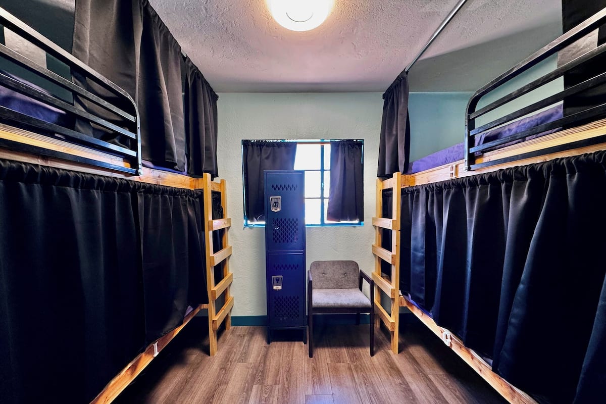 Een studentenkamer met stapelbedden, een kleine stoel en een raam met gordijnen.