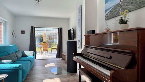 A living room with a piano, a teal sofa, and a dining area with chairs and a table. - Apartment Wilhelmine ground floor - winter garden, quiet and central in Aurich Ostfriesland (Aurich)