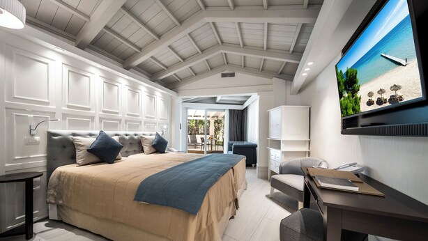 A bedroom with a large bed, a desk, a chair, and a TV displaying a beach scene. - Forte Village Resort Villa Del Parco & Spa (Pula)
