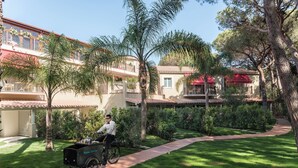 A person riding a bicycle with a cart labeled "Room Service" through a well-maintained courtyard with palm trees and a pathway. - Forte Village Resort Villa Del Parco & Spa (Pula)
