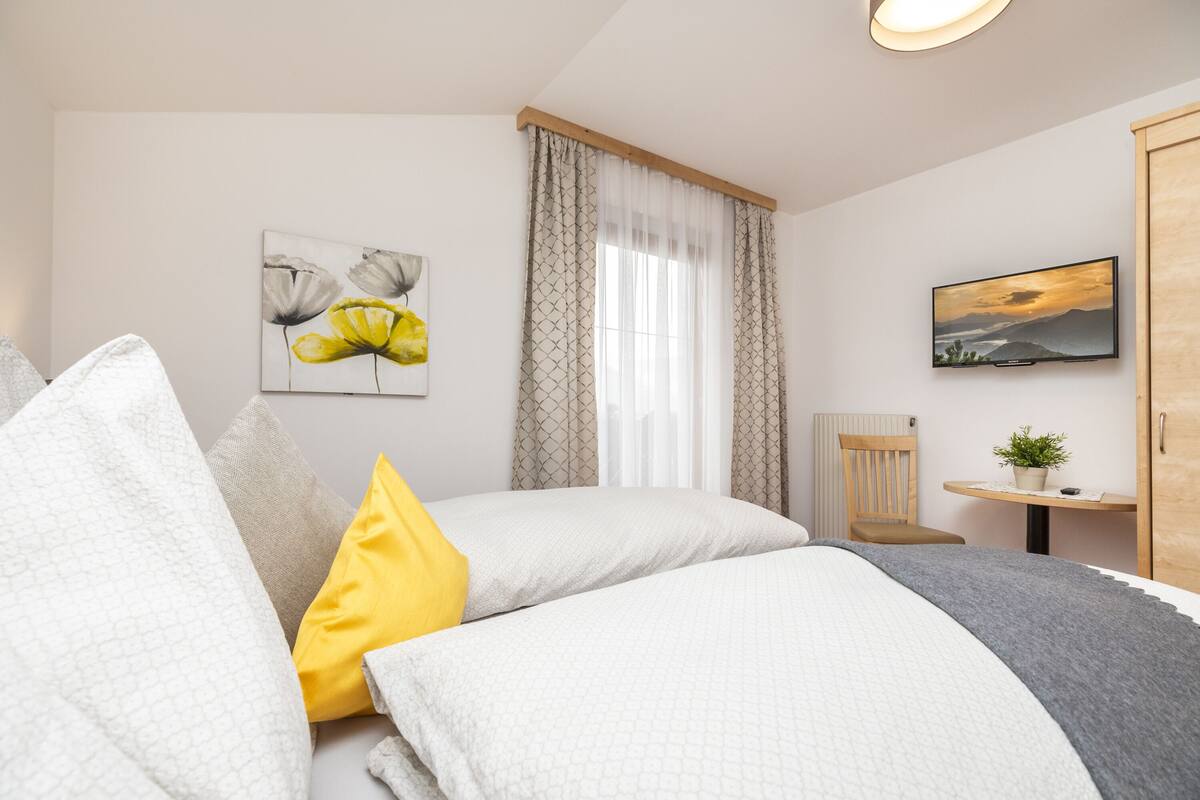 Un dormitorio con cama, almohadas, ventana con cortinas, un cuadro, televisor, una silla, una mesita con planta y un armario.