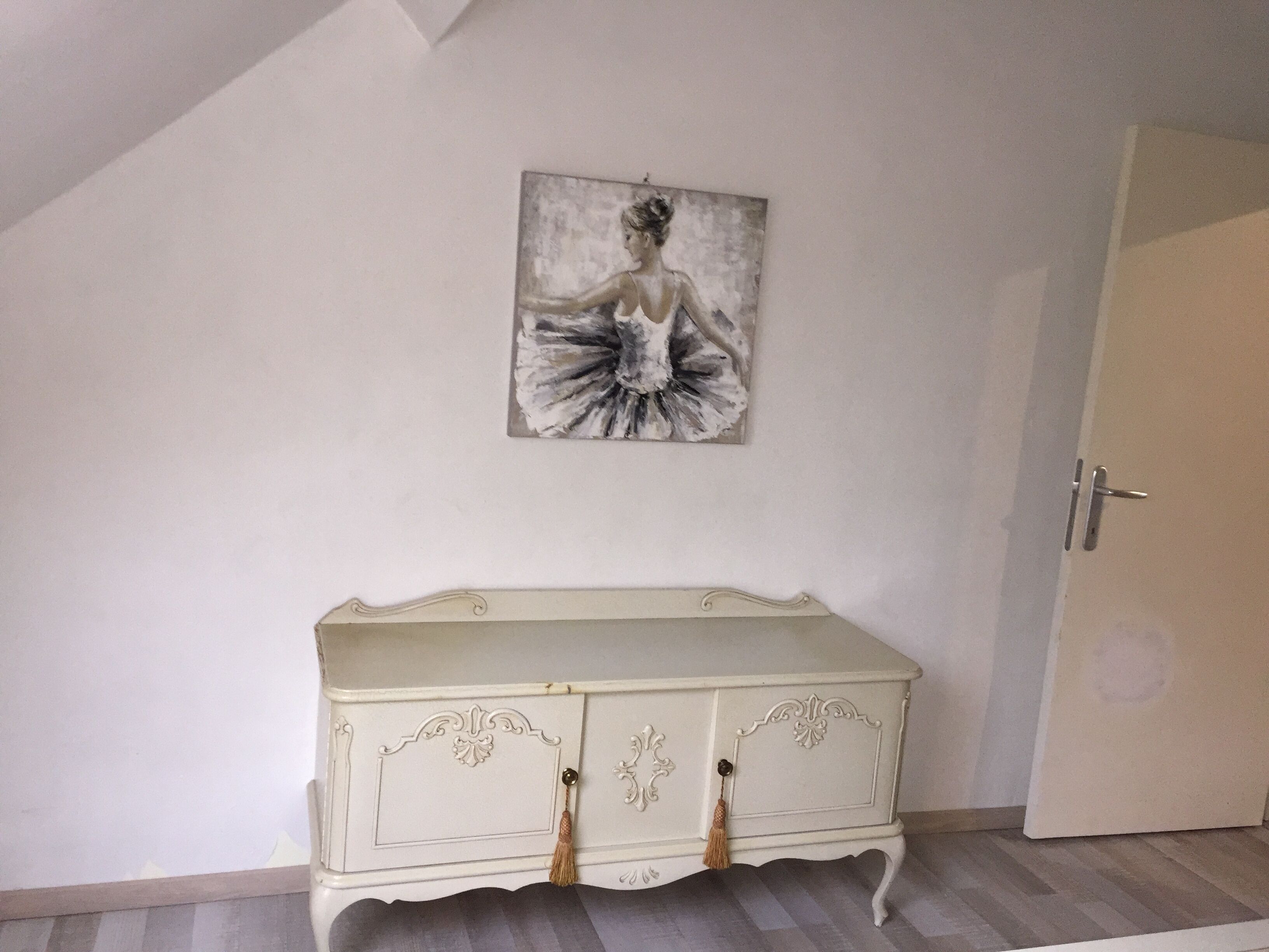 A room with a white wall, a white cabinet with decorative handles, and a painting of a woman in a dress.