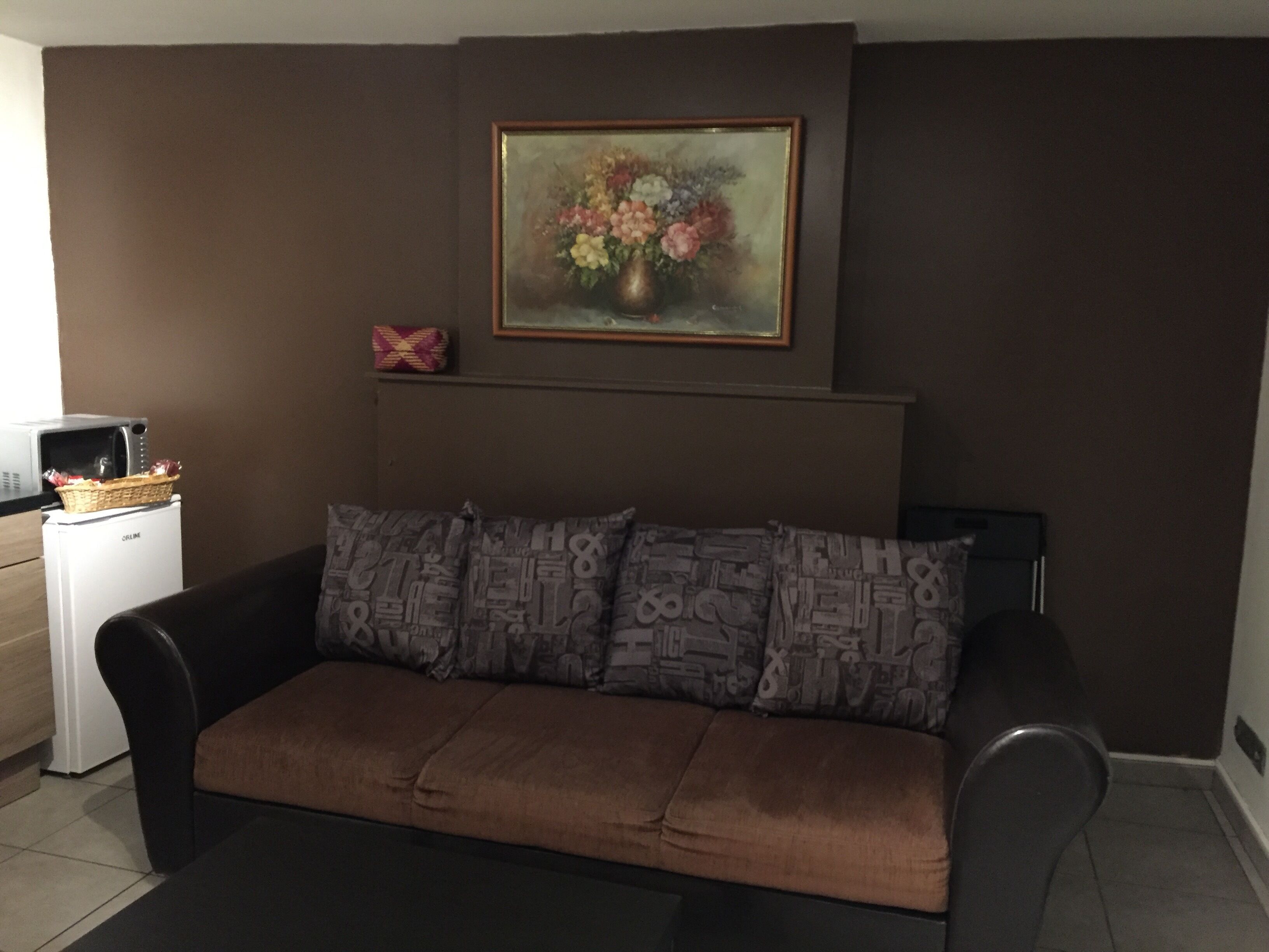 A living room with a brown sofa, a framed floral painting above it, and a small white appliance on the left.