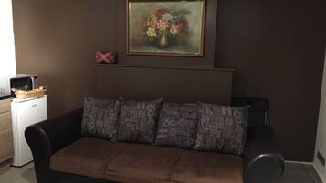 A living room with a brown sofa, a framed floral painting above it, and a small white appliance on the left.