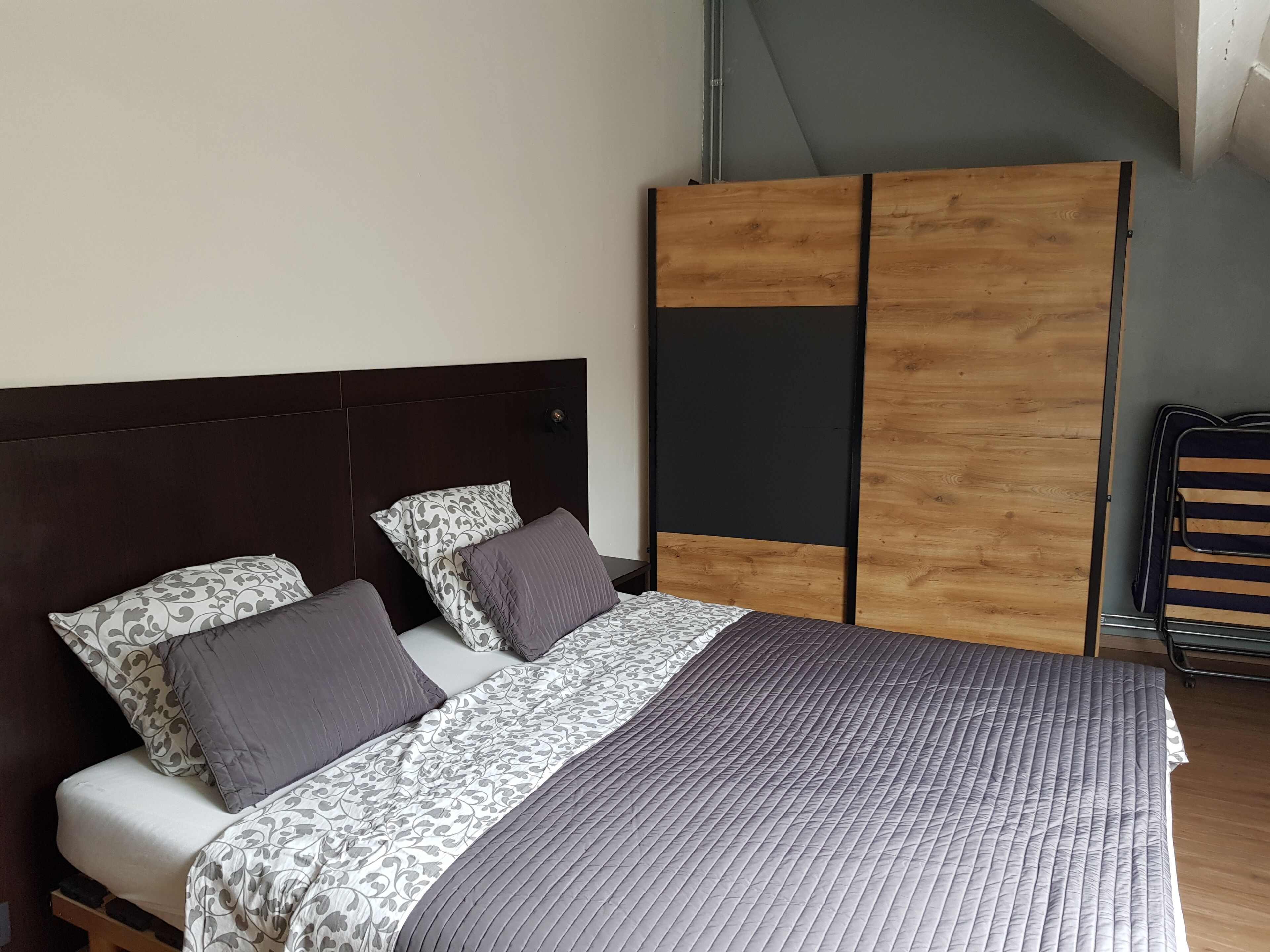 Een net opgemaakt bed met een bedspreide en kussens met een patroon, een donker hoofdbord en een houten garderobe met een schuifdeur.