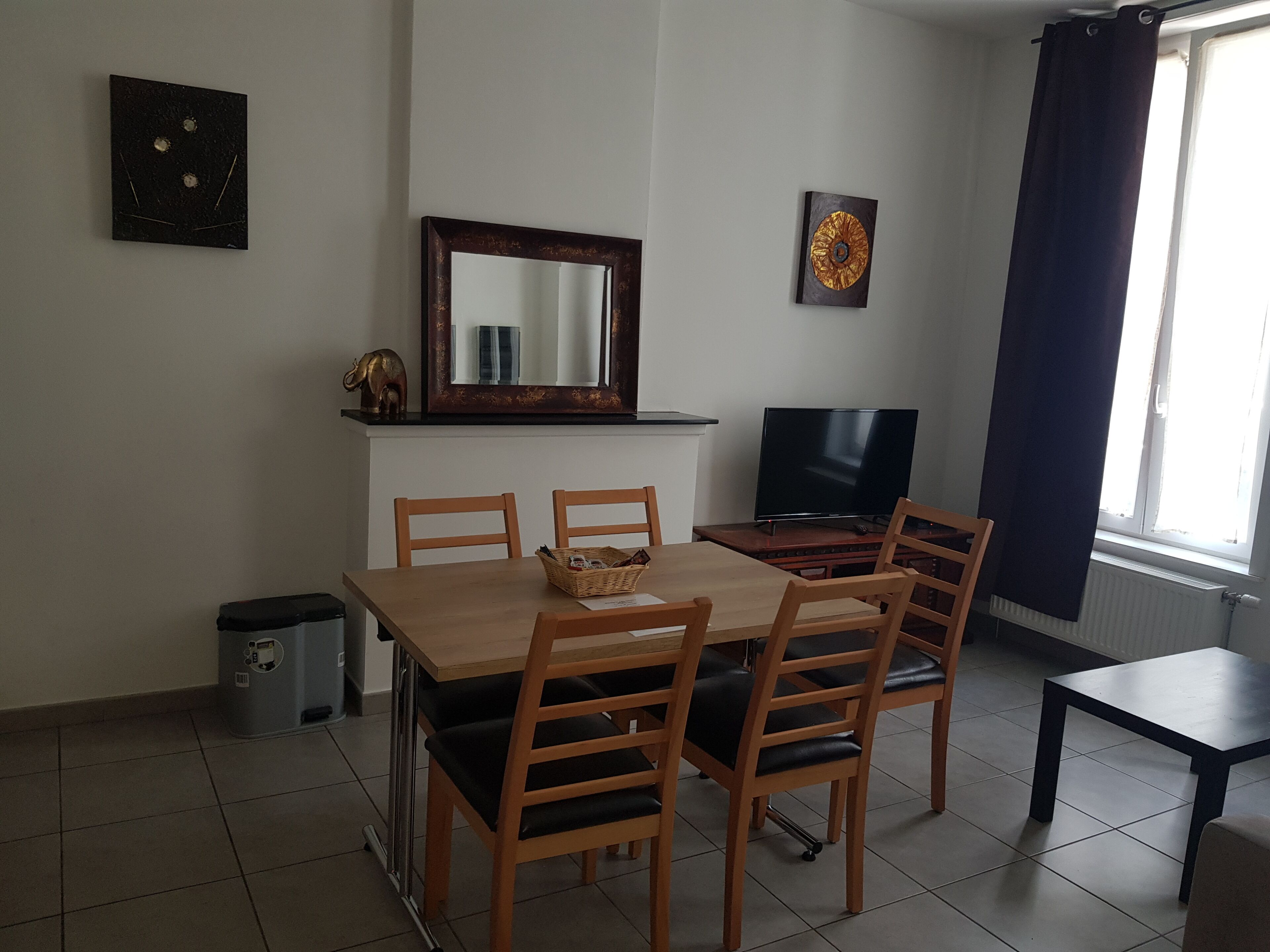 A dining area with a wooden table, four chairs, a television, and a small trash bin.