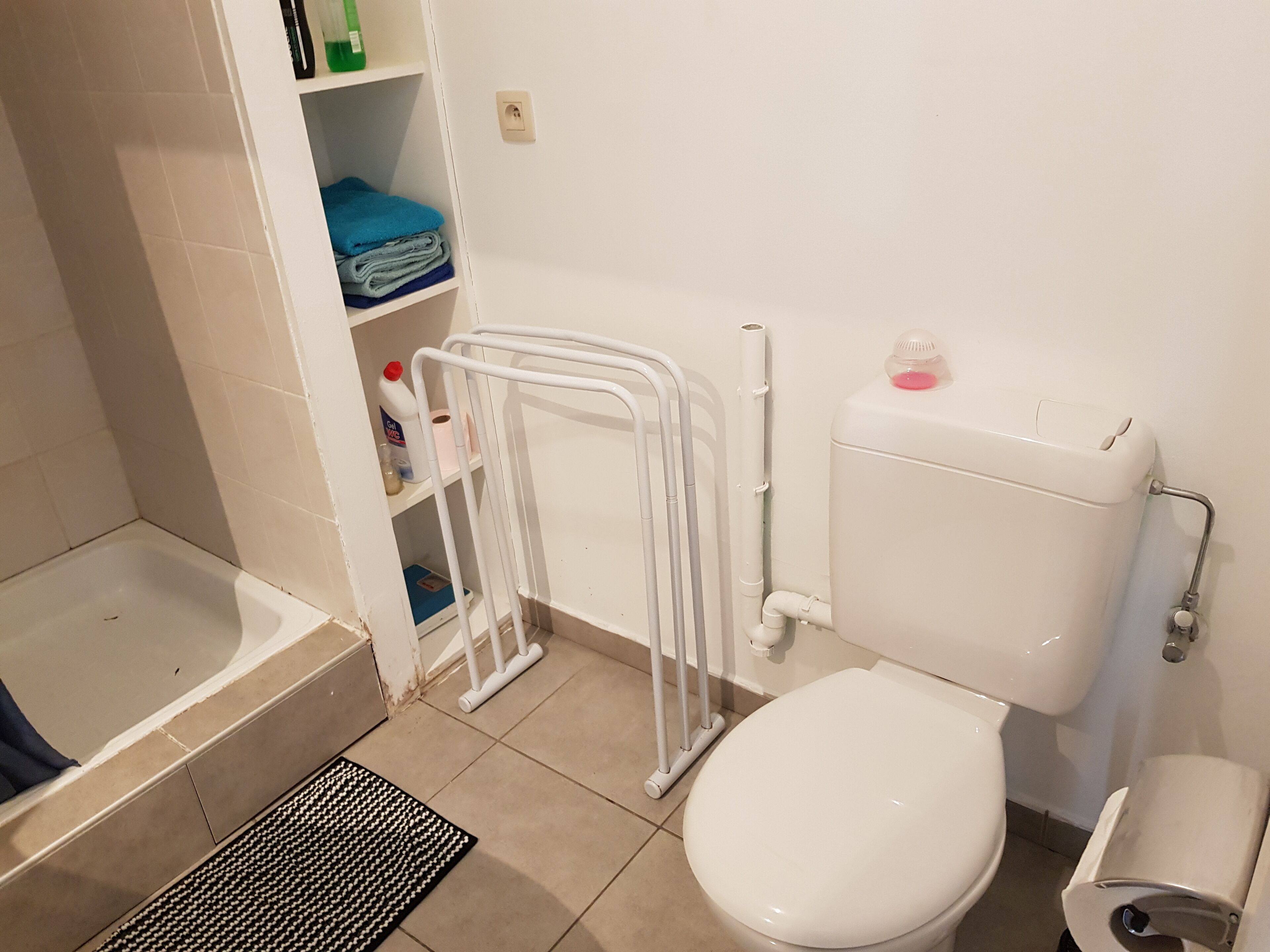 Ein Badezimmer mit Toilette, einem weißen Heizkörper, einem Regal mit gefalteten Handtüchern und einer Badewanne.