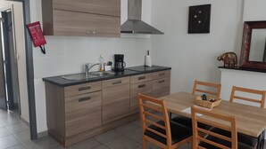 A modern kitchen with wooden cabinets, a stainless steel sink, and a coffee maker. There is a dining table with chairs and a small decorative elephant figure. - Appartements bon Secours (Peruwelz)