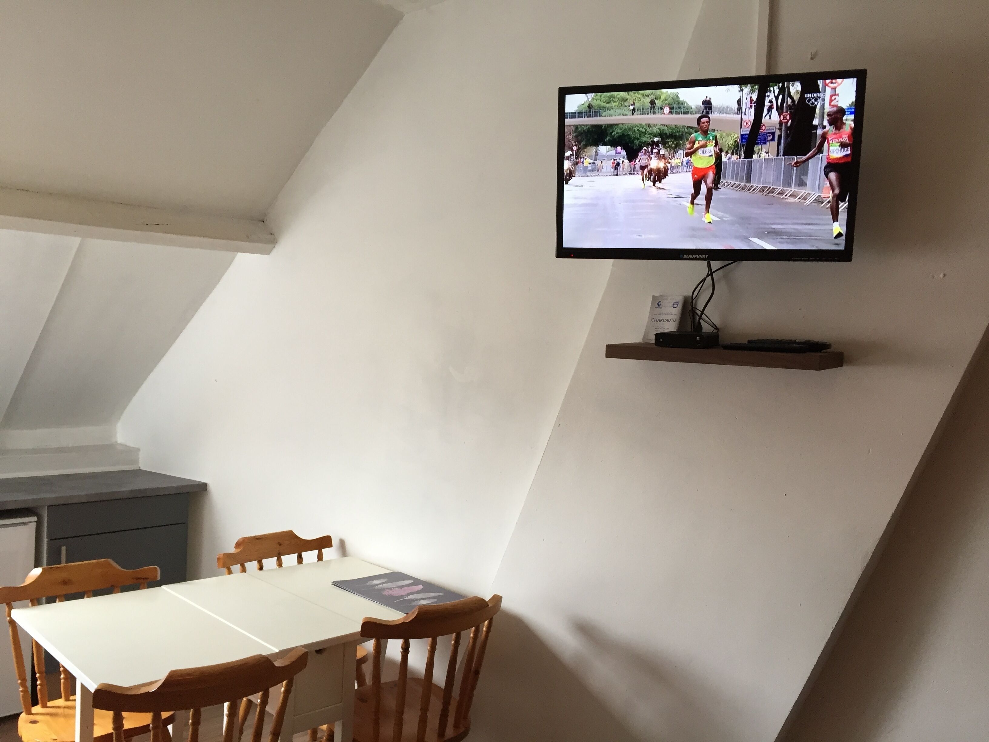 Un téléviseur fixé à un mur incliné, diffusant un marathon, une table blanche avec des chaises et une petite étagère située sous le téléviseur.