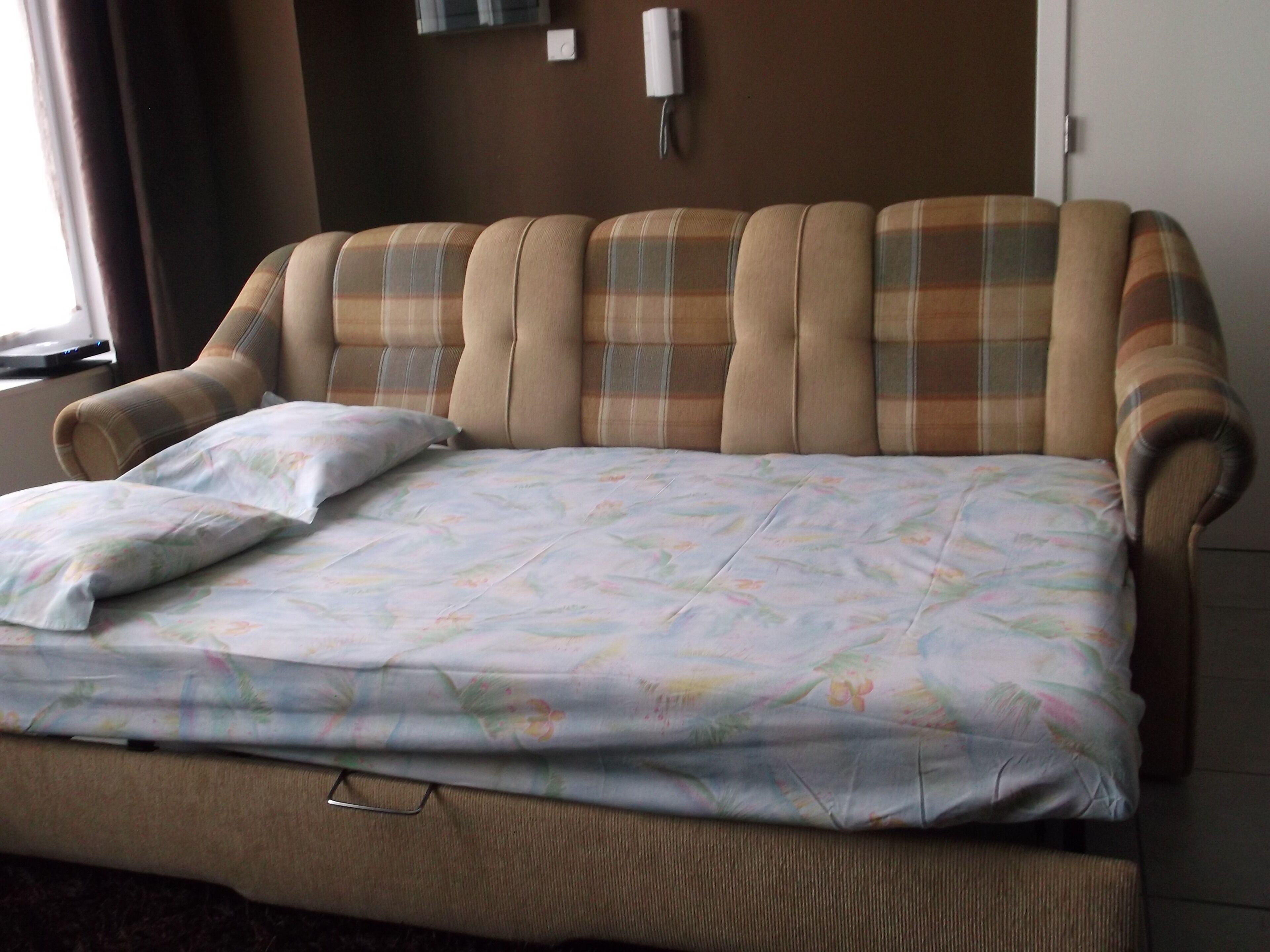 A bed with a floral bedspread and a pillow, a headboard with a plaid pattern, and a wall-mounted light fixture.