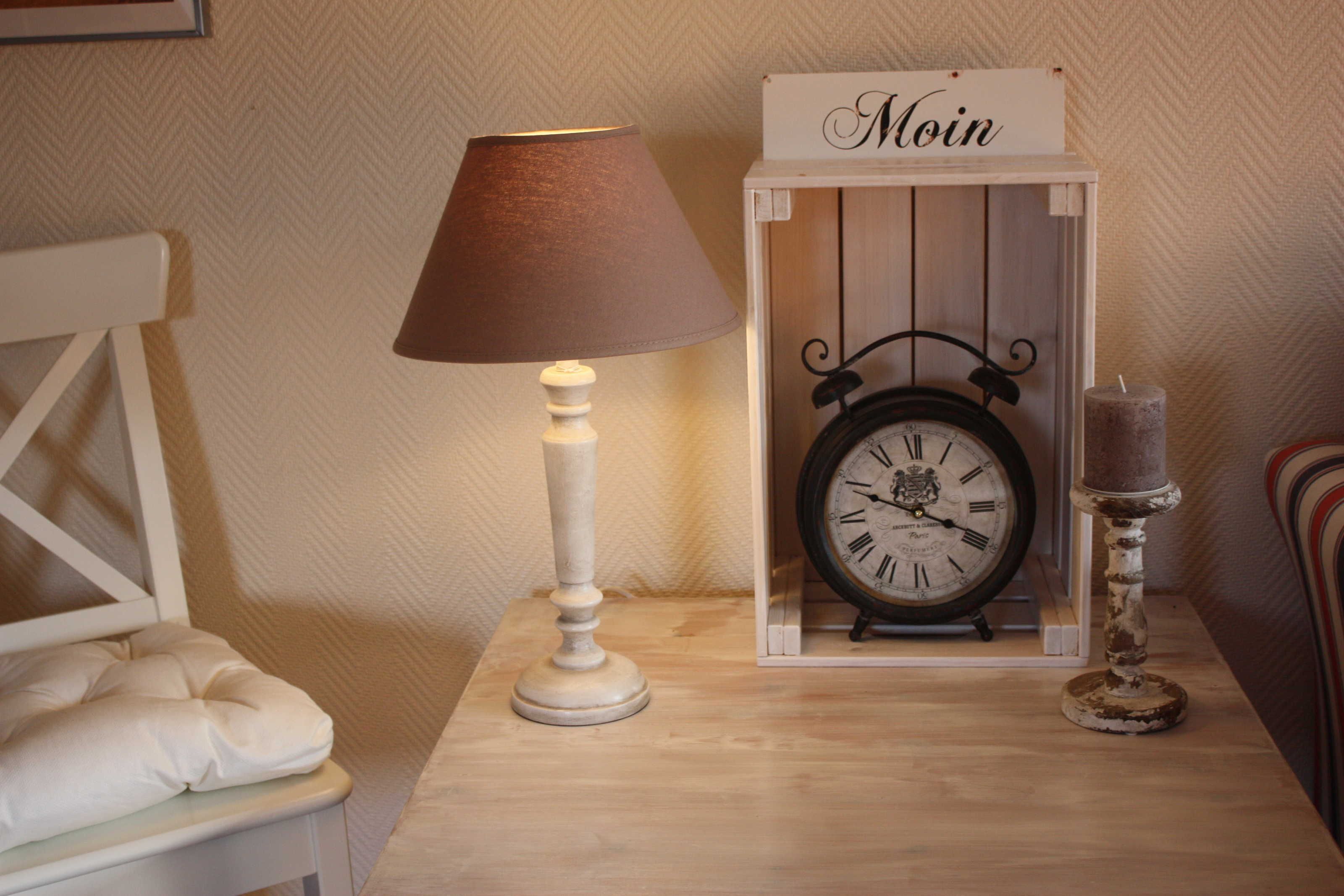 Een nachtkastje met een witte lamp, een klok, een kaars en een bordje met de tekst 'Moin'.