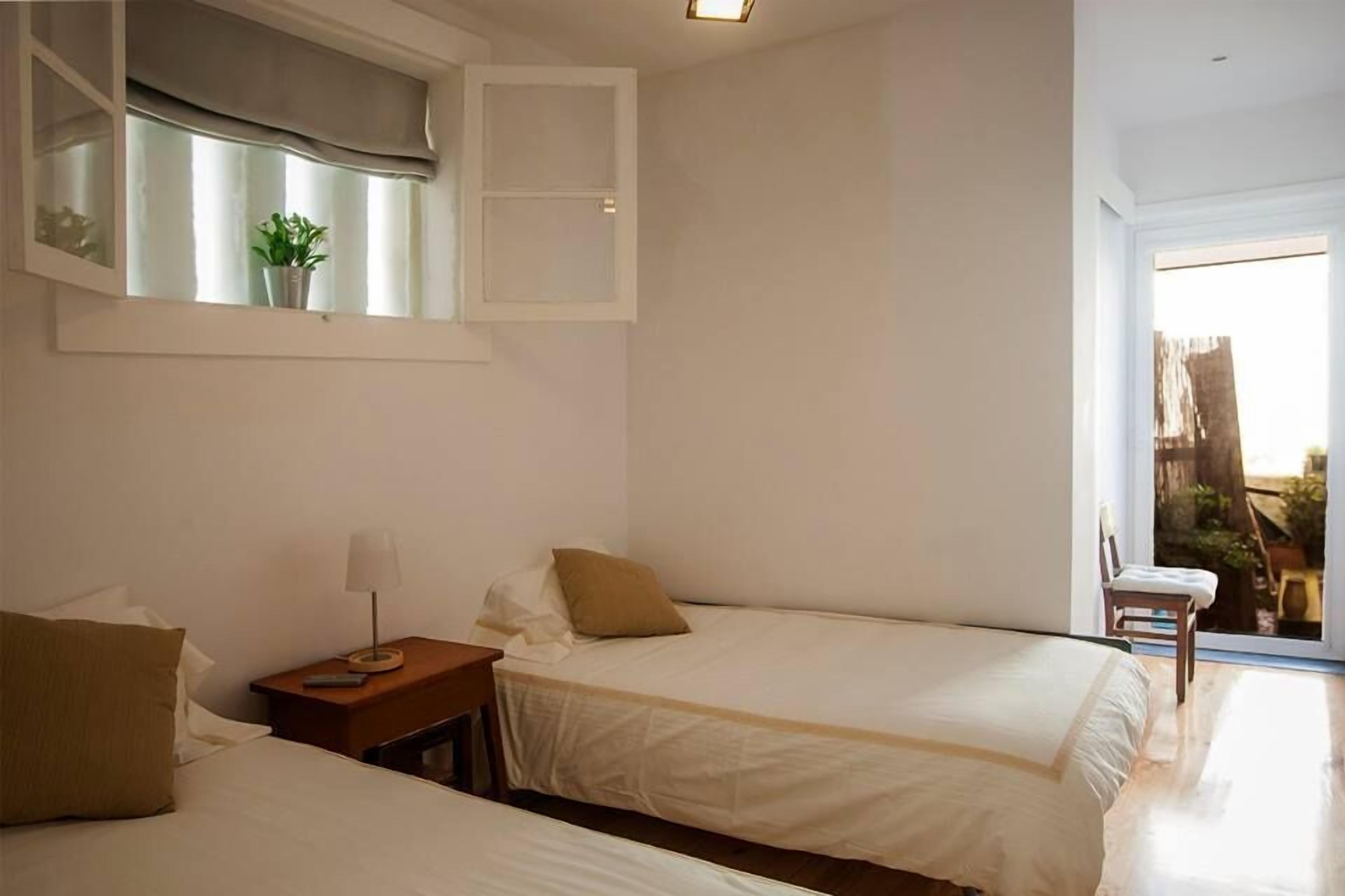 A hotel room with two beds, a small table with a lamp, a window with a plant, and a view of the outside.