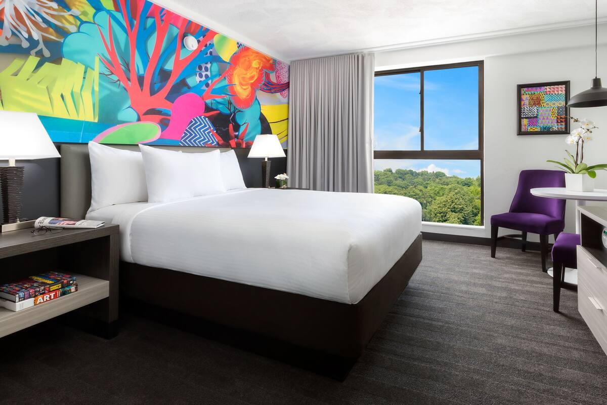 Une chambre d’hôtel avec un grand lit, un bureau avec une chaise, une télévision, une lampe de bureau, une bibliothèque, une fenêtre donnant sur un paysage verdoyant et une peinture abstraite aux couleurs vives accrochée au mur.
