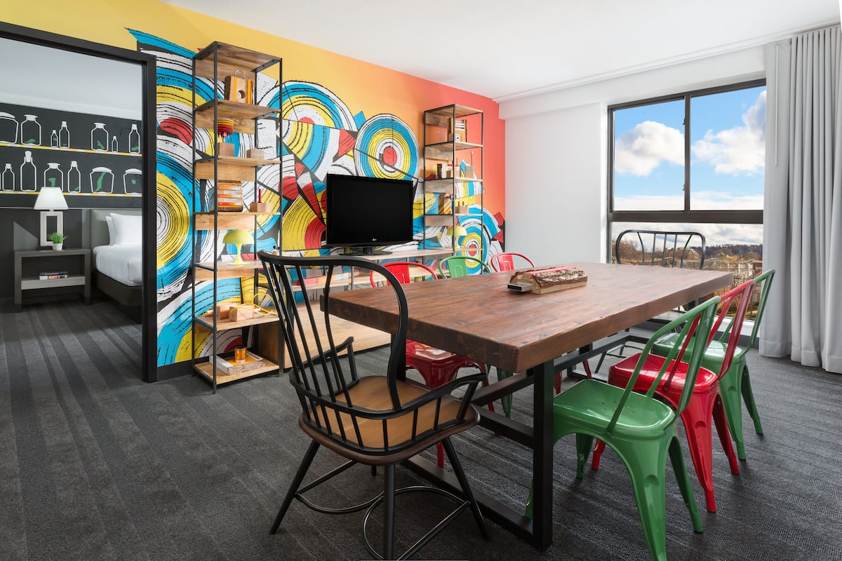 Une salle de réunion moderne, dotée d’une table en bois, de chaises colorées, d’un téléviseur noir et d’une fresque murale abstraite aux couleurs vives.
