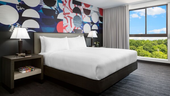 A hotel room with a large bed, two bedside tables, a nightstand, a bookshelf, and a view of greenery through the window.