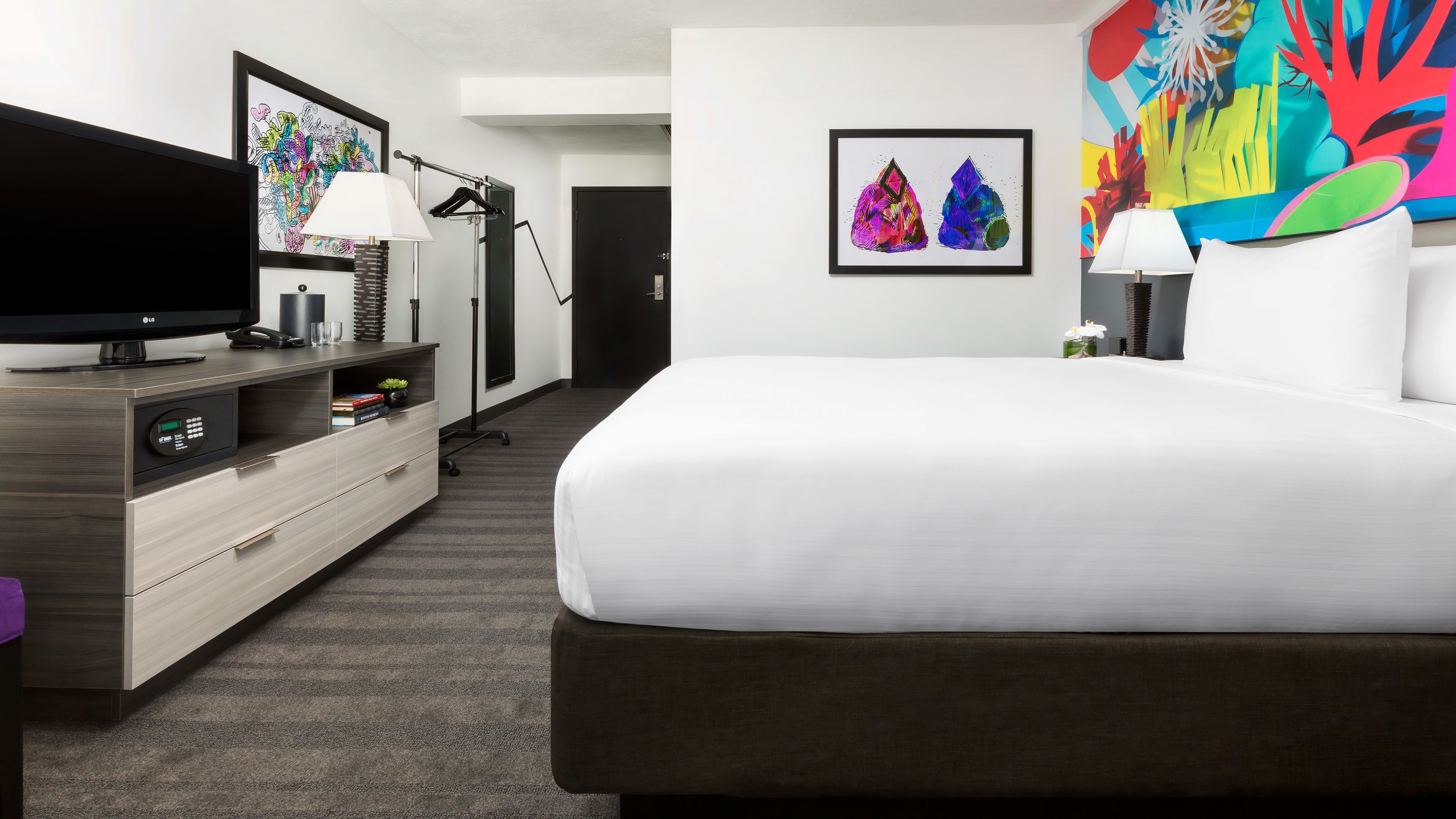 Ein Hotelzimmer mit einem Bett, einem Fernseher, einem Schreibtisch mit Lampe und Kunstwerken an den Wänden.