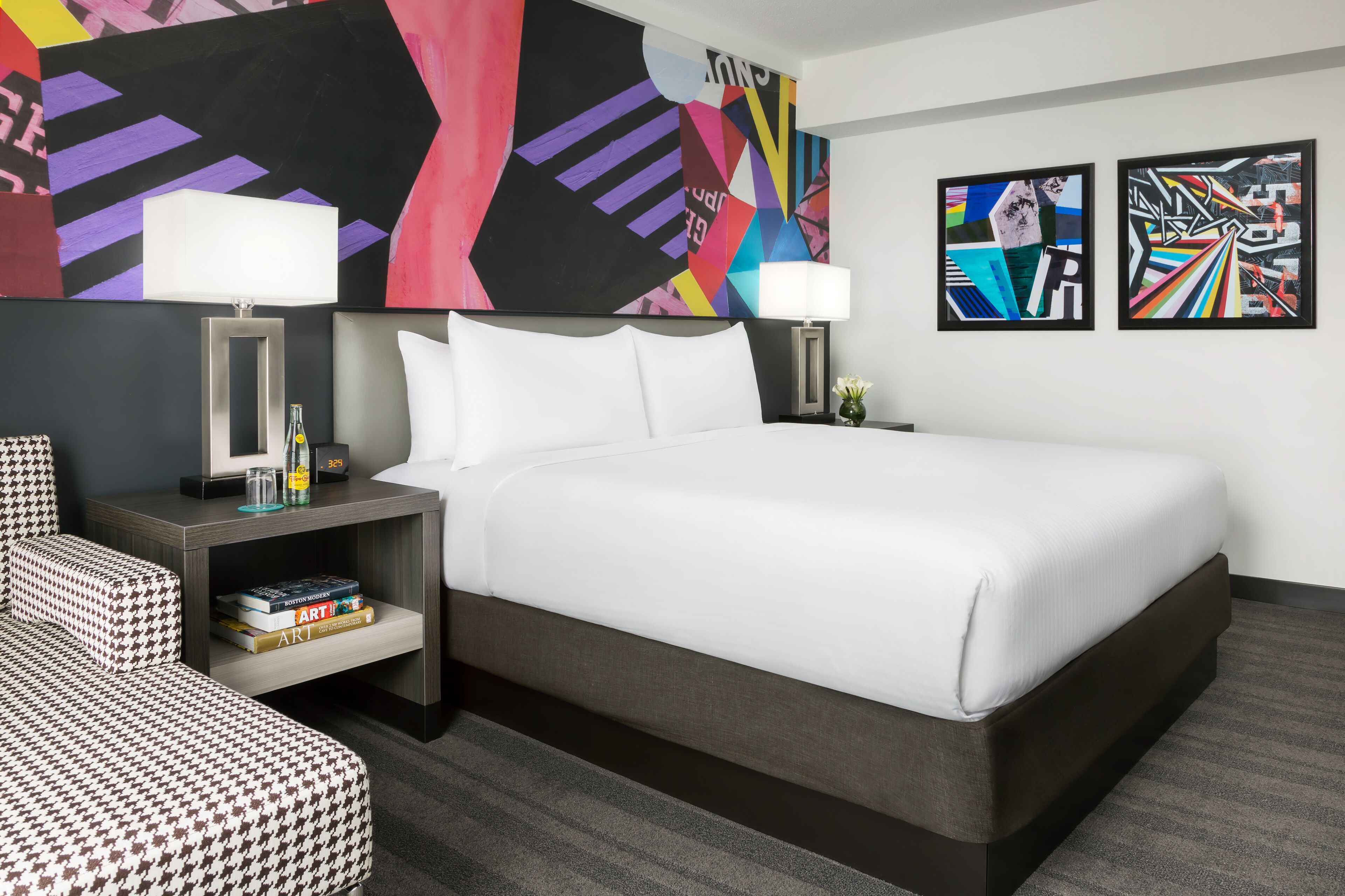 Habitación de hotel moderna con una cama grande, dos mesitas de noche con lámparas, un estante con libros y obras de arte abstracto en las paredes.