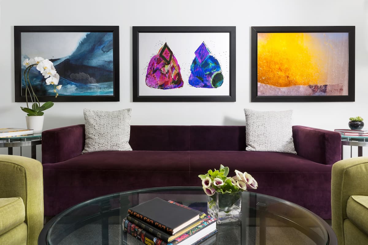 Un salon avec un canapé de couleur violet foncé, une table basse en verre sur laquelle sont posés des livres, et trois œuvres d’art encadrées accrochées au mur.