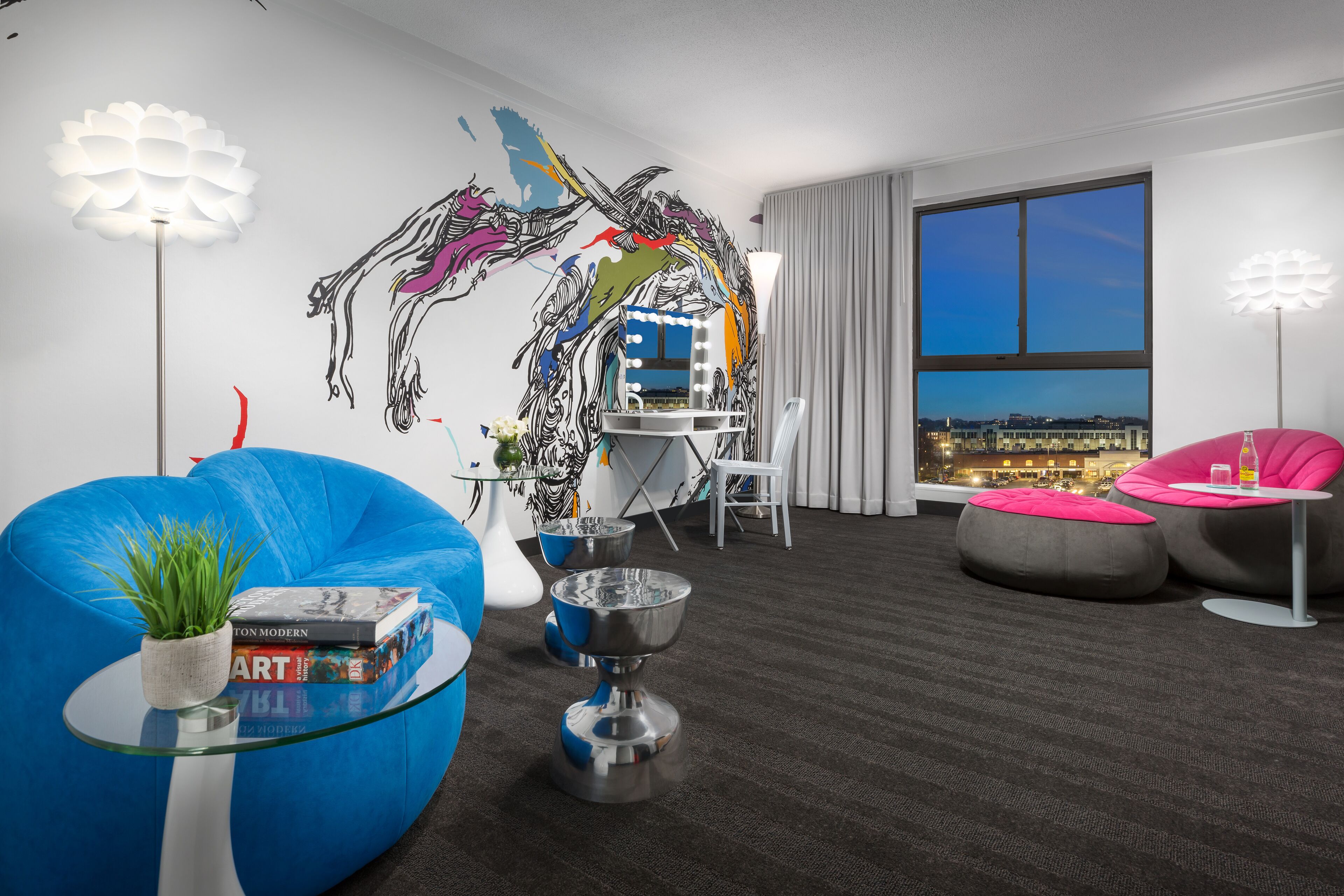 A modern hotel room with a large window, a mural, a blue bean bag, a glass coffee table, and a pink chair.